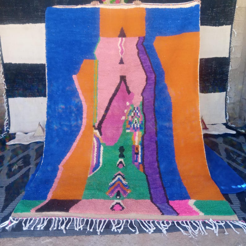 May include: A vibrant, abstract rug with a colorful design. The rug features a combination of blue, orange, pink, green, and purple hues. It has a fringed edge and a geometric pattern, adding a touch of modern art to any space.