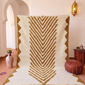 K&ouml;nnte beinhalten: Ein cremefarbener, rechteckiger Teppich mit einem geometrischen Chevron-Muster in Braunt&ouml;nen. Der Teppich hat einen Fransenrand und eine Zickzack-Bord&uuml;re. Der Teppich wird in einem Raum mit einer dekorativen Laterne und einem Lederhocker ausgestellt.
