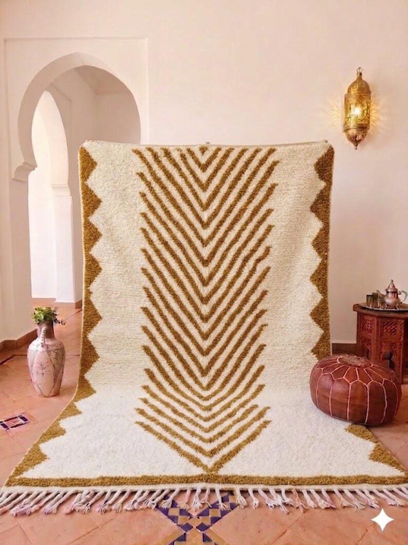 K&ouml;nnte beinhalten: Ein cremefarbener, rechteckiger Teppich mit einem geometrischen Chevron-Muster in Braunt&ouml;nen. Der Teppich hat einen Fransenrand und eine Zickzack-Bord&uuml;re. Der Teppich wird in einem Raum mit einer dekorativen Laterne und einem Lederhocker ausgestellt.