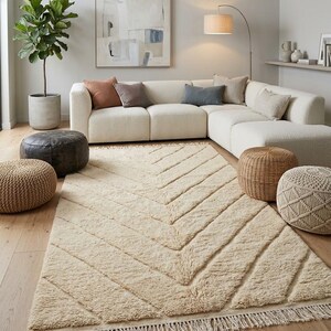 Op de afbeelding: Een rechthoekig vloerkleed in een lichtbeige kleur met een verhoogd visgraatpatroon, een hoogpolige textuur en franjes aan de korte zijden. Het kleed ligt op een lichte houten vloer voor een witte hoekbank.
