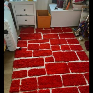 May include: A rectangular rug with a brick pattern. The rug features red rectangular shapes separated by white spaces, creating a brick wall design. The rug is placed on a wooden floor.
