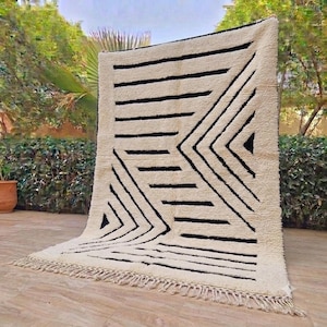 May include: A rectangular area rug featuring a thick, cream-colored pile with a bold, black geometric pattern consisting of horizontal lines and chevron-like shapes. The rug is finished with a knotted fringe along the bottom edge.