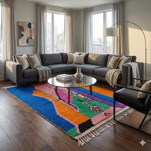 May include: A modern living room featuring a large gray sectional sofa, a round glass coffee table, and a vibrant, colorful area rug. The rug has a bold design with blue, orange, pink, and green hues. The room is well-lit with natural light.