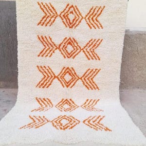 K&ouml;nnte beinhalten: Cremefarbener, rechteckiger Teppich mit einem geometrischen Muster in Oranget&ouml;nen. Das Design zeigt sich wiederholende Diamant- und Pfeilformen. Der Teppich hat eine strukturierte Oberfl&auml;che und einen Fransenbesatz am unteren Rand.