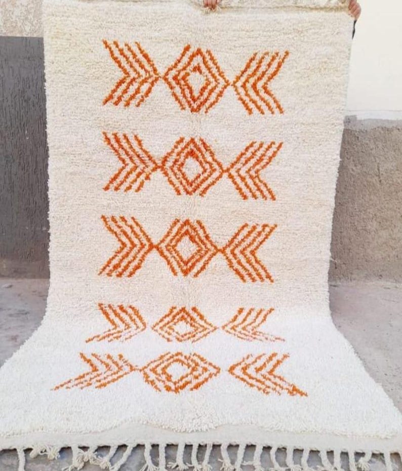 K&ouml;nnte beinhalten: Cremefarbener, rechteckiger Teppich mit einem geometrischen Muster in Oranget&ouml;nen. Das Design zeigt sich wiederholende Diamant- und Pfeilformen. Der Teppich hat eine strukturierte Oberfl&auml;che und einen Fransenbesatz am unteren Rand.