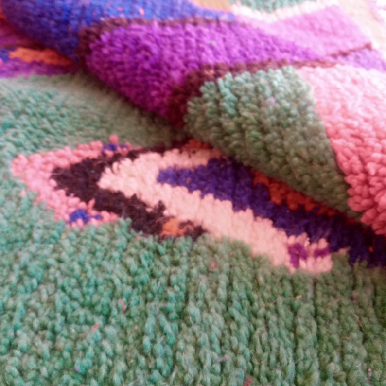 May include: Close-up of a colorful, textured rug with a geometric pattern. The rug features a mix of green, purple, pink, and blue hues. The design includes abstract shapes and a soft, plush texture, suggesting a handmade or artisanal quality.