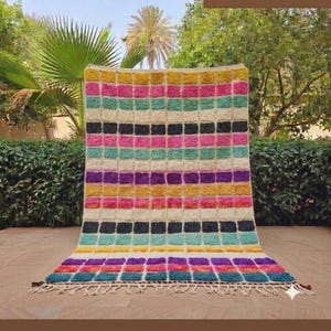 May include: A rectangular area rug featuring a grid pattern of thick, plush, multi-coloured horizontal stripes in shades of yellow, pink, teal, black, and purple against a cream-coloured background, finished with a knotted fringe along the bottom edge.