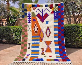 Puede incluir: Una alfombra rectangular con base de color crema y un patrón geométrico multicolor. El diseño incluye formas naranjas, azules, rojas, verdes y moradas, con un borde a cuadros y flecos blancos en el borde inferior.