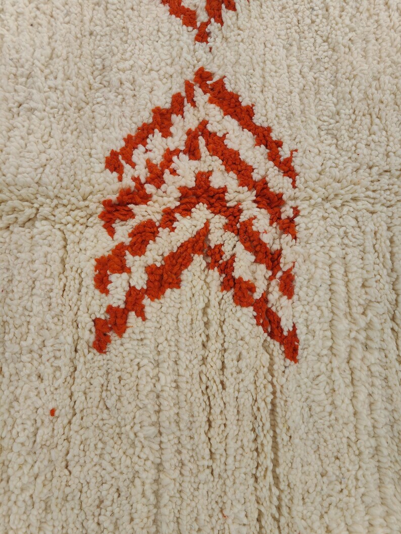 K&ouml;nnte beinhalten: Cremefarbener Teppich mit strukturierter Oberfl&auml;che und einem kr&auml;ftigen, geometrischen Design in Orange- und Wei&szlig;t&ouml;nen. Das Muster zeigt eine Reihe von pfeilartigen Formen, die einen auff&auml;lligen visuellen Kontrast erzeugen. Geeignet f&uuml;r Wohn- und Schlafzimmer.
