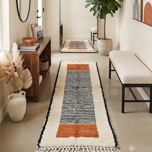 May include: A long rectangular runner rug featuring a cream-colored base with a central panel of thin horizontal black stripes. The rug includes two solid burnt orange rectangular blocks at each end and is finished with white fringe tassels.