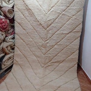 Op de afbeelding: Een groot, rechthoekig, cr&egrave;mekleurig vloerkleed met een dikke, hoogpolige textuur. Het oppervlak is versierd met een herhalend visgraatpatroon dat wordt gevormd door verzonken lijnen in de stof. De onderrand is afgewerkt met franjes.
