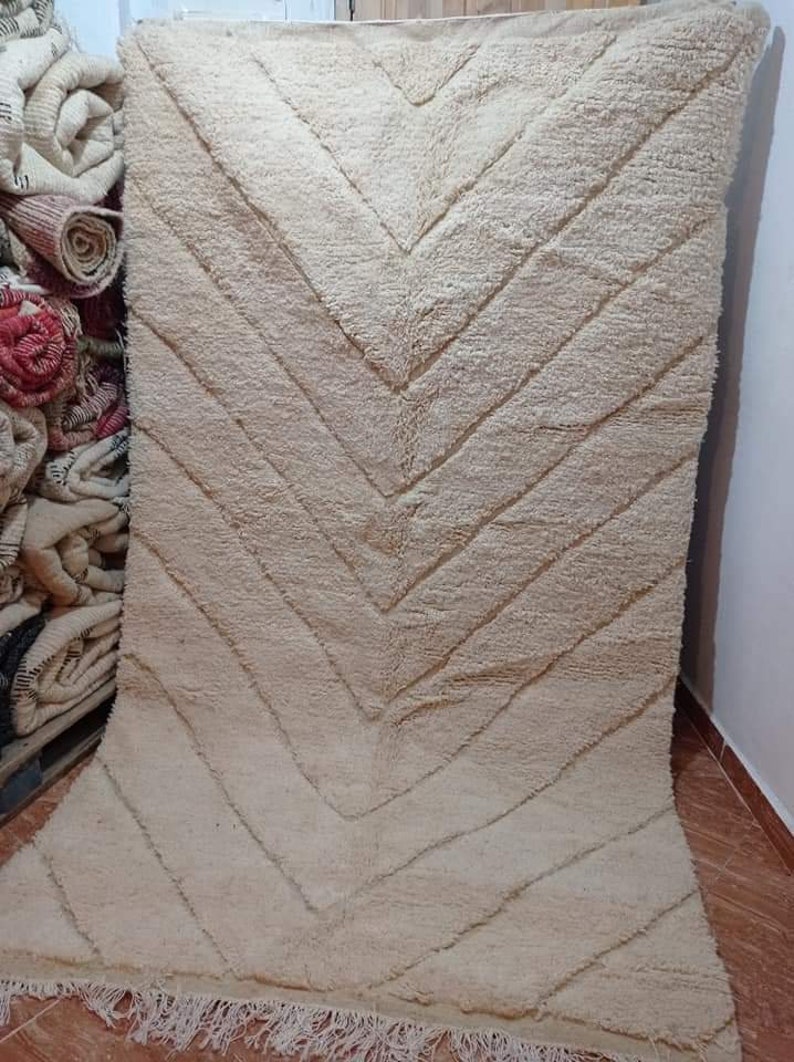 Op de afbeelding: Een groot, rechthoekig, cr&egrave;mekleurig vloerkleed met een dikke, hoogpolige textuur. Het oppervlak is versierd met een herhalend visgraatpatroon dat wordt gevormd door verzonken lijnen in de stof. De onderrand is afgewerkt met franjes.