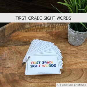 Puede incluir: Un conjunto de tarjetas de memoria con las palabras "First Grade Sight Words" impresas en el frente. Las tarjetas están dispuestas en forma de abanico y tienen un anillo de encuadernación plateado.