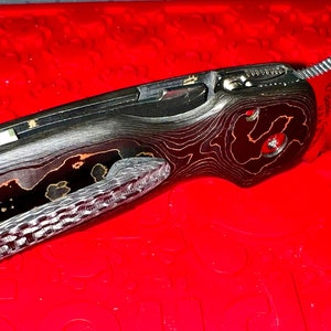 May include: A black and orange patterned pocket knife with a silver blade and a hole in the handle. The knife is closed and lying on a red surface.