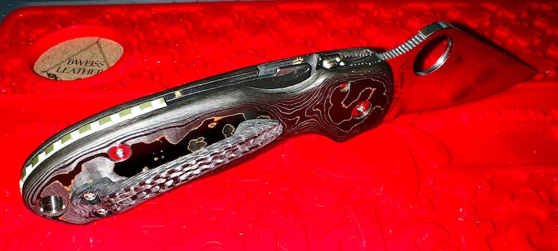 May include: A black and orange patterned pocket knife with a silver blade and a hole in the handle. The knife is closed and lying on a red surface.