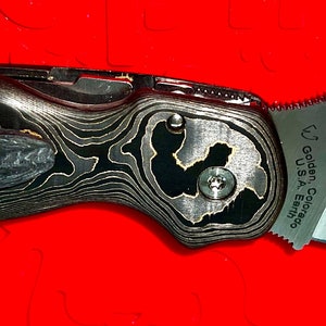 May include: A black and gold pocket knife with a partially open blade. The blade has a serrated edge and the handle has a unique wood grain pattern. The knife has the text "Spyderco USA" and "LF" on the blade.