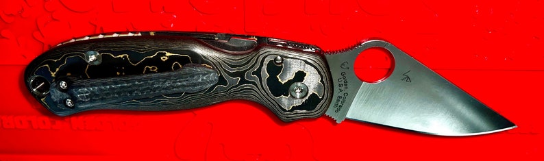 May include: A black and gold pocket knife with a partially open blade. The blade has a serrated edge and the handle has a unique wood grain pattern. The knife has the text "Spyderco USA" and "LF" on the blade.