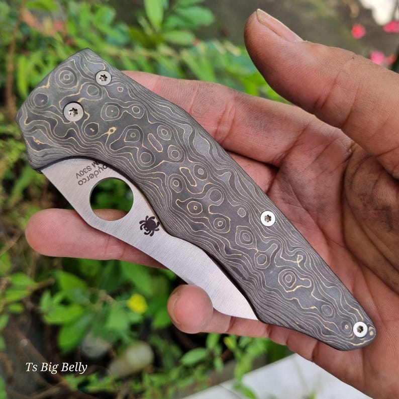 May include: A Spyderco knife with a black and gold patterned handle and a silver blade. The knife is closed and the handle has the text "Spyderco" and "S30V" on it.