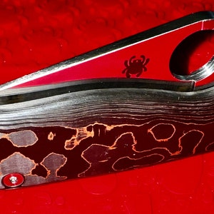 May include: A black and red pocket knife with a spider logo on the blade. The handle is made of a patterned metal.
