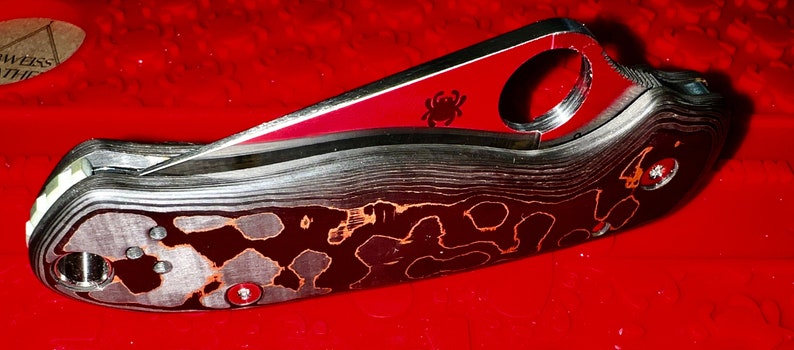 May include: A black and red pocket knife with a spider logo on the blade. The handle is made of a patterned metal.