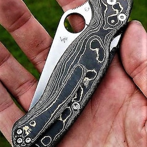May include: A black and gold pocket knife with a patterned handle. The knife has a small hole in the handle and a partially visible blade.