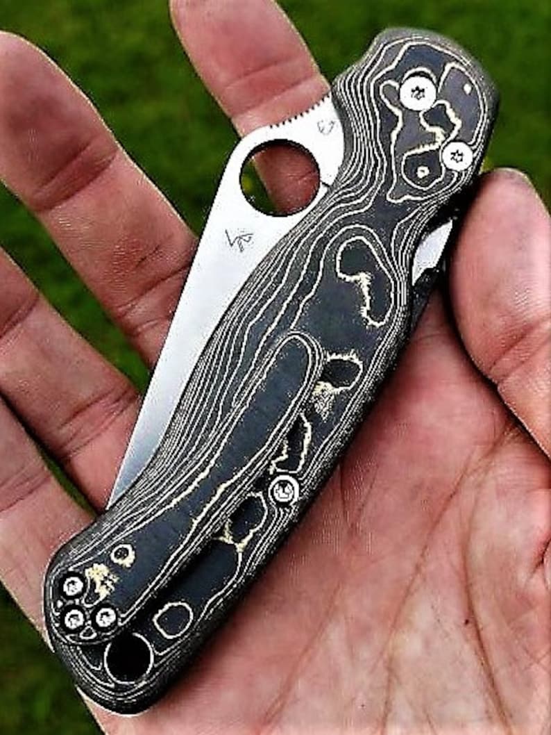 May include: A black and gold pocket knife with a patterned handle. The knife has a small hole in the handle and a partially visible blade.