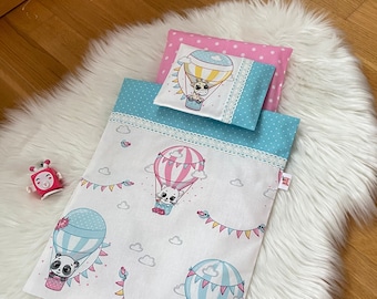 May include: A white and blue children's bedding set with a hot air balloon theme. The bedding features a white background with pink and blue hot air balloons, each with a cute animal inside. The bedding also has a blue and white polka dot pillow with a pink hot air balloon and a giraffe inside.