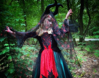 May include: A child dressed as a fairy tale character wearing a black and red dress with a black lace overlay and a black crown. The dress has a green and black tulle skirt. The child is standing in a forest setting.