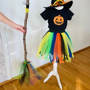 May include: A black witch costume for kids with a tulle skirt in rainbow colors, a black t-shirt with a pumpkin graphic, and a black witch hat with orange and yellow accents. The costume also includes a broomstick made from a branch with yellow and green tulle.