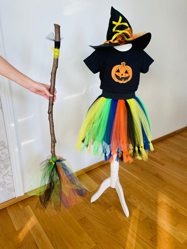 May include: A black witch costume for kids with a tulle skirt in rainbow colors, a black t-shirt with a pumpkin graphic, and a black witch hat with orange and yellow accents. The costume also includes a broomstick made from a branch with yellow and green tulle.