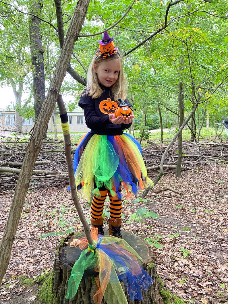 May include: A young girl dressed in a Halloween costume with a black shirt, a rainbow tulle skirt, orange and black striped tights, and black boots. She is holding a small orange pumpkin with a witch's hat and a spider on top. She is wearing a headband with two small orange pumpkins with witch's hats on top. The girl is standing on a tree stump in a wooded area.