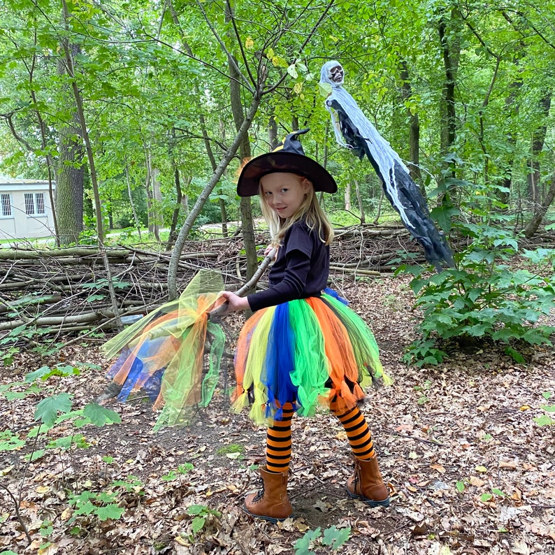 May include: A young girl dressed as a witch for Halloween. She is wearing a black witch hat with a green and orange striped tulle skirt. She is holding a broom and smiling.