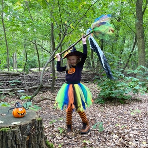 May include: A young girl dressed as a witch for Halloween, wearing a black shirt with a pumpkin design, a colorful tutu skirt, and a witch's hat. She is holding a broom and smiling.