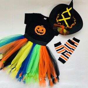 May include: A Halloween costume set for a child, featuring a black witch hat with a yellow ribbon, a black t-shirt with a pumpkin graphic, a rainbow tulle skirt with tassels, and black and white striped leg warmers.