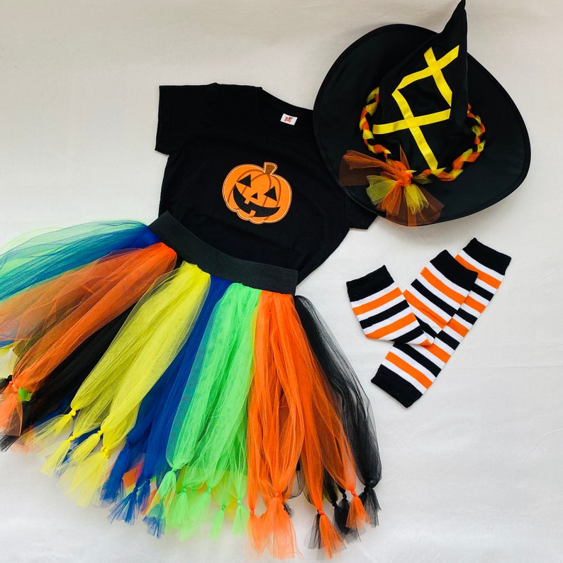 May include: A Halloween costume set for a child, featuring a black witch hat with a yellow ribbon, a black t-shirt with a pumpkin graphic, a rainbow tulle skirt with tassels, and black and white striped leg warmers.