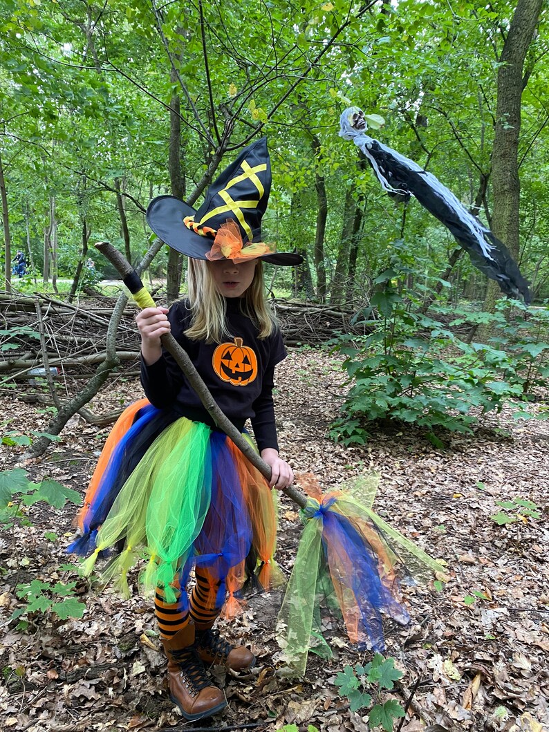 May include: A young girl dressed as a witch for Halloween. She is wearing a black witch hat with yellow and orange accents, a black shirt with a pumpkin design, a colorful tulle skirt, and striped tights. She is holding a broomstick made of a branch with colorful tulle streamers.