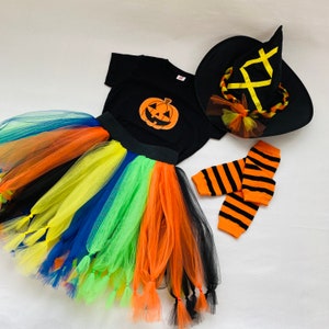 May include: A Halloween costume set for a child, featuring a black shirt with an orange pumpkin applique, a rainbow tulle skirt with black and orange accents, a black witch hat with yellow and orange accents, and orange and black striped leg warmers.
