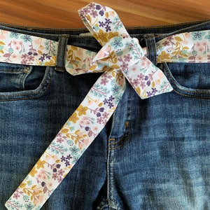 Peut inclure: Une ceinture en tissu blanc avec un motif floral dans les tons rose, violet et jaune. La ceinture est nouée en un nœud autour d'un jean bleu.