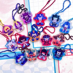 May include: A selection of colourful acrylic keychains, each depicting a cartoon robot character. The keychains, in various colours, are attached to ball chains. The background is a patterned surface.