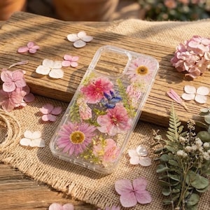 May include: Clear phone case with embedded dried flowers in shades of pink, purple, and yellow. The case is surrounded by scattered flower petals and sprigs of greenery on a wooden surface.