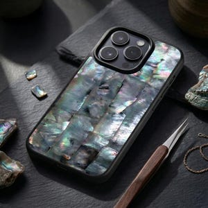 May include: A black smartphone case featuring a mosaic inlay of iridescent abalone shell pieces. The shell surface displays a spectrum of shimmering green, blue, and purple tones. The case has a black frame with a triple-lens camera cutout.