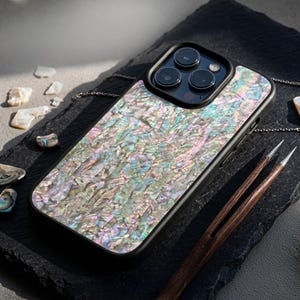 Può includere: Custodia per smartphone nera con un design cangiante e iridescente in madreperla. La custodia ha un bordo nero e un ritaglio per la fotocamera. L'immagine mostra anche un paio di pinzette di legno e piccole conchiglie.