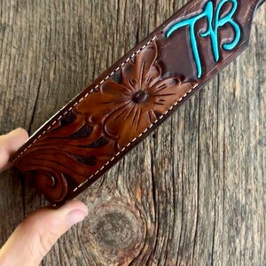May include: A brown leather strap featuring a hand-tooled floral pattern and the letters TB in bright turquoise. The strap is finished with white stitching along the edges and a silver-toned metal rivet at the top corner of the leather piece.