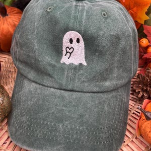 May include: A washed-out dark green baseball cap featuring an embroidered white ghost graphic on the front panel. The ghost has two black oval eyes and a small black heart shape on its chest. The cap has a curved brim and a button on top.