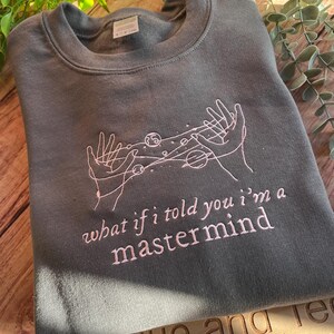 May include: A dark gray sweatshirt with a pink embroidered design of two hands holding a constellation of planets and stars. The text "what if i told you i'm a mastermind" is also embroidered in pink.