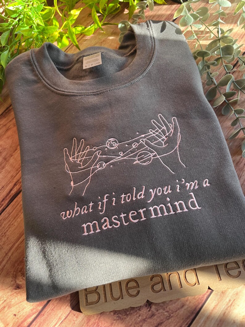 May include: A dark gray sweatshirt with a pink embroidered design of two hands holding a constellation of planets and stars. The text "what if i told you i'm a mastermind" is also embroidered in pink.