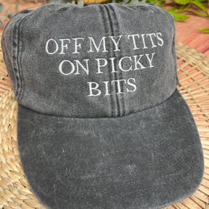 May include: A charcoal grey baseball cap featuring the white embroidered text "OFF MY TITS ON PICKY BITS". The cap has a curved peak and a worn, vintage appearance. The hat is made of a soft, durable material.
