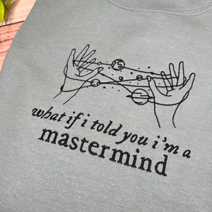 May include: A light blue sweatshirt with a black embroidered design of two hands holding a galaxy with planets. The text "what if i told you i'm a mastermind" is embroidered below the design.
