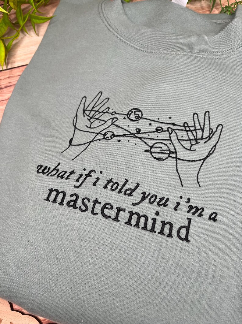 May include: A light blue sweatshirt with a black embroidered design of two hands holding a galaxy with planets. The text "what if i told you i'm a mastermind" is embroidered below the design.