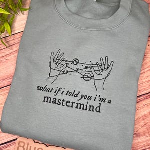 May include: A light blue sweatshirt with a black graphic of hands holding planets and stars. The text "what if i told you i'm a mastermind" is printed below the graphic.